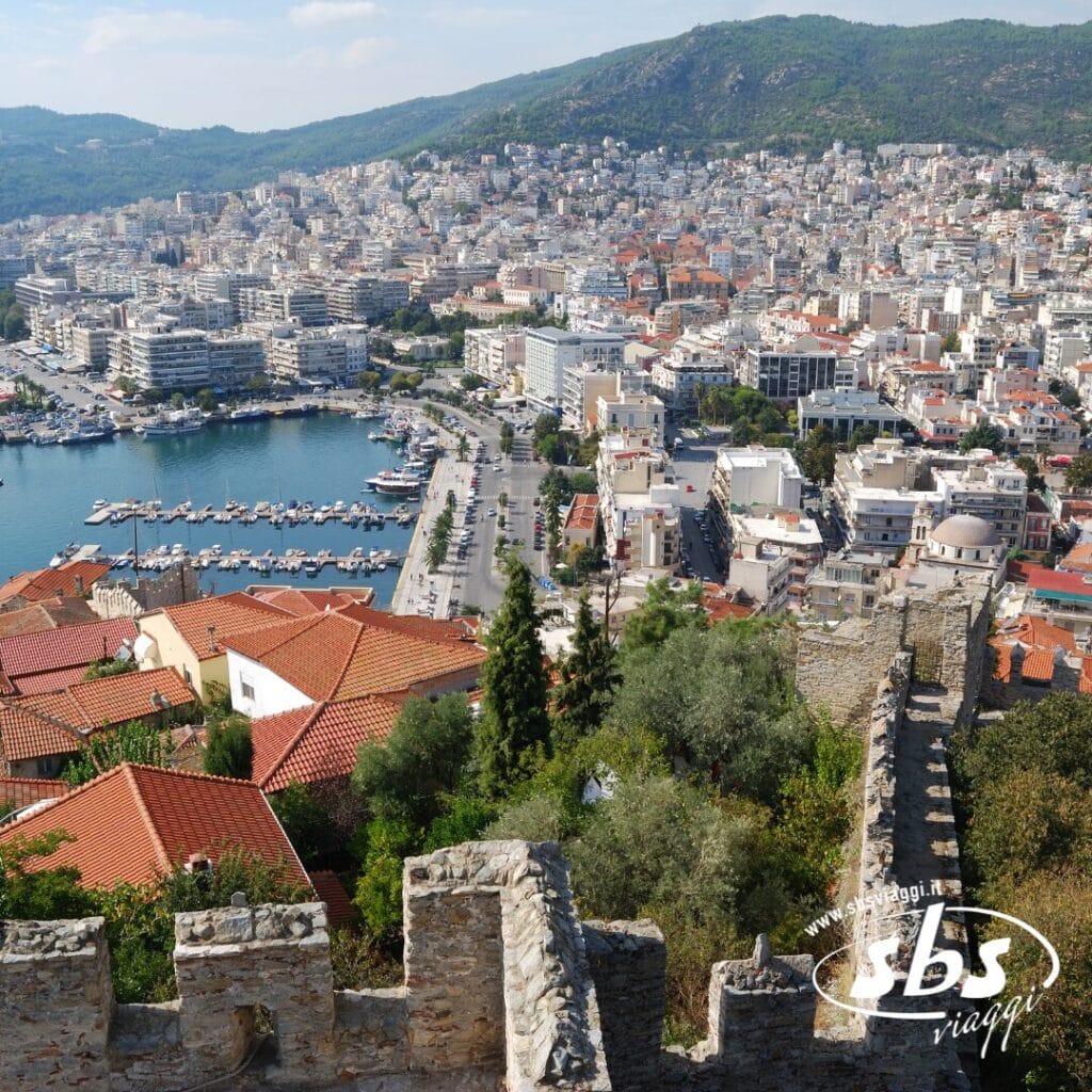 Una città costiera con un porto turistico, edifici dai tetti rossi e uno storico muro di pietra in primo piano, sullo sfondo di colline e un denso sviluppo urbano: scopri Salonicco durante il tuo prossimo tour nelle Terre di Alessandro Magno.