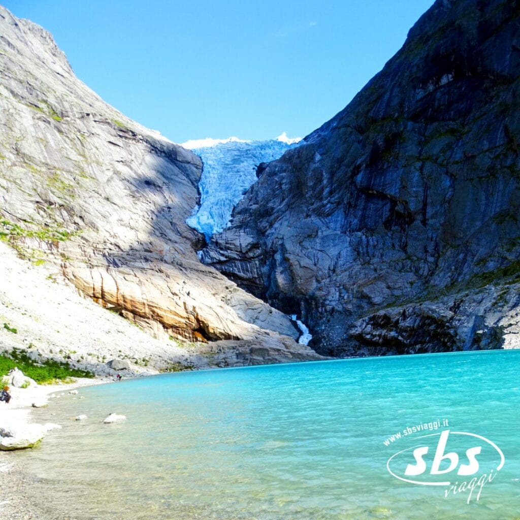 Un lago di un azzurro brillante si trova sotto un ghiacciaio incastonato tra ripide montagne rocciose e sotto un cielo azzurro e limpido, creando uno spettacolo naturale degno dei Fiordi.
