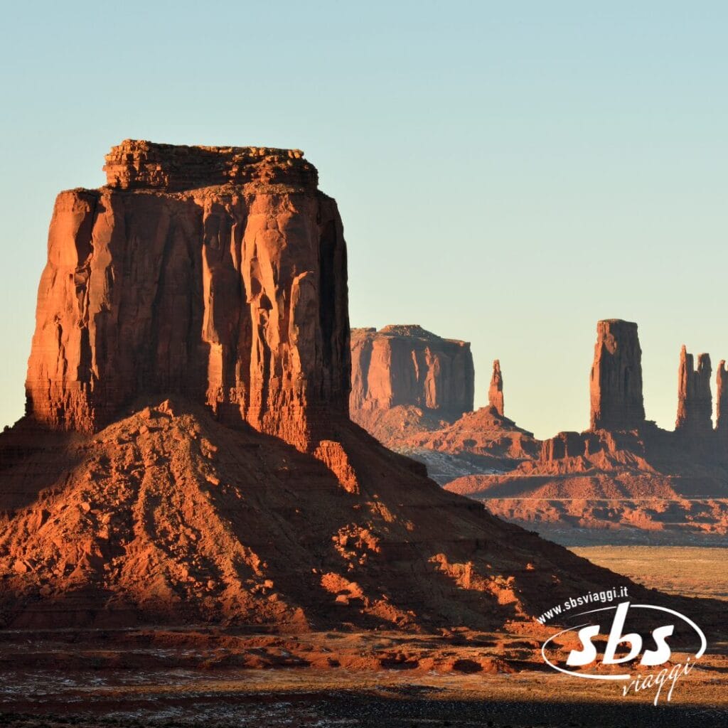 Le colline e le altopiani della Monument Valley sono illuminate dalla luce del sole, proiettando lunghe ombre sul paesaggio desertico; nell'angolo in basso a destra appare il logo della compagnia di viaggi Explorer 1924.
