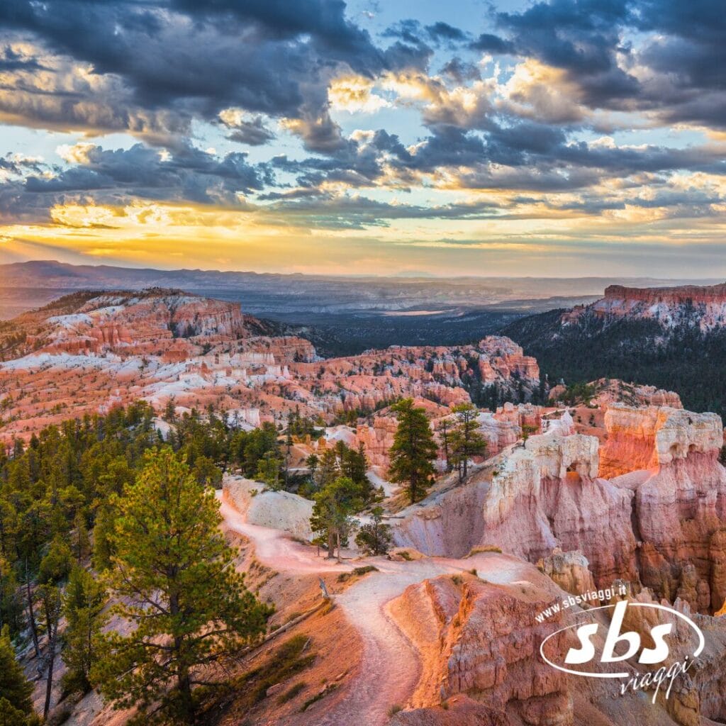 Un sentiero tortuoso si snoda attraverso le formazioni rocciose color arancio del Bryce Canyon, sotto un cielo parzialmente nuvoloso al tramonto; gli alberi punteggiano il paesaggio, evocando una scena lontana da Los Angeles o dal Clear Sky del 1924.