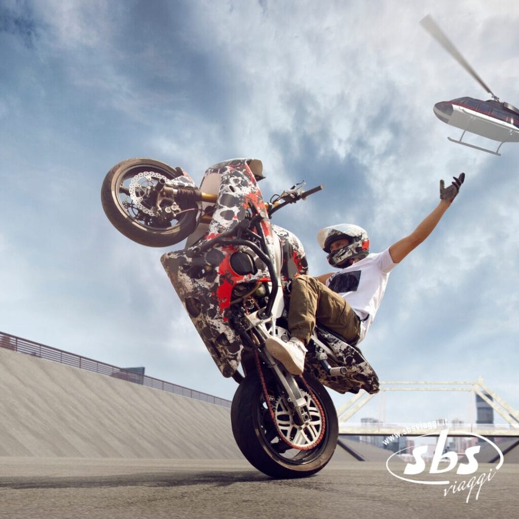 Un motociclista esegue un'audace impennata su una rampa con una mano alzata, mentre un elicottero vola sullo sfondo. L'immagine, con il logo