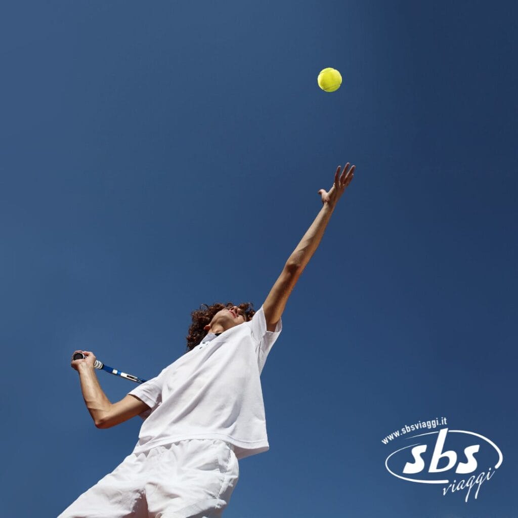 Una persona in abbigliamento sportivo bianco si prepara a colpire una palla da tennis contro un cielo azzurro e terso, tenendo la racchetta sopra la testa. Il logo