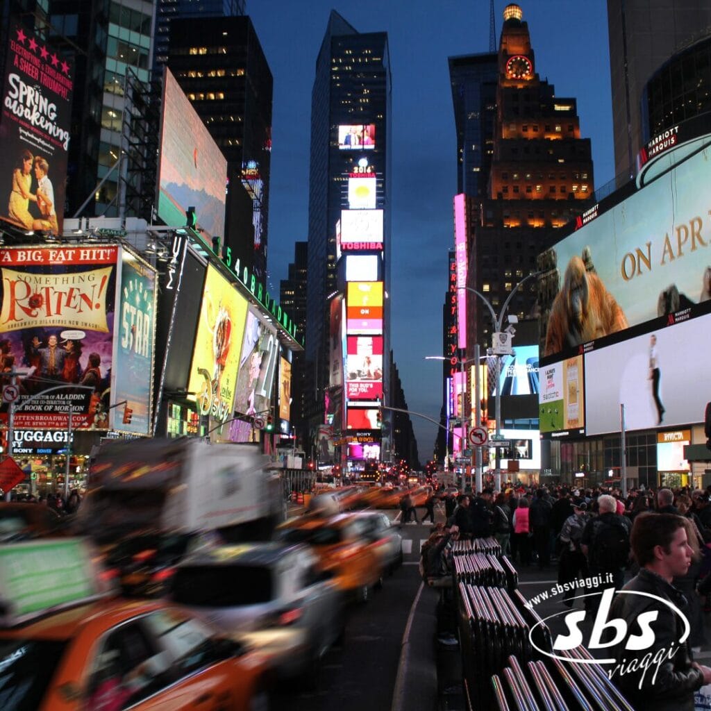 Times Square affollata al tramonto con cartelloni pubblicitari elettronici luminosi, folla sul marciapiede, taxi che attraversano la strada e tifosi che indossano magliette NBA diretti al Madison Square Garden.