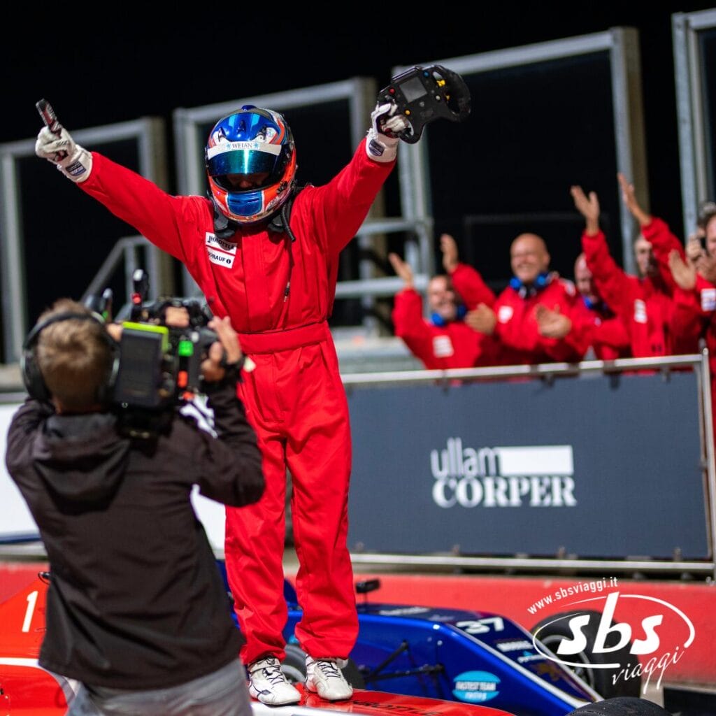 Un pilota di Formula 1 con indosso una tuta rossa e un casco è in piedi su un'auto con le braccia alzate, mentre un operatore di ripresa cattura le emozioni in pista mentre le persone in uniforme rossa applaudono sullo sfondo.