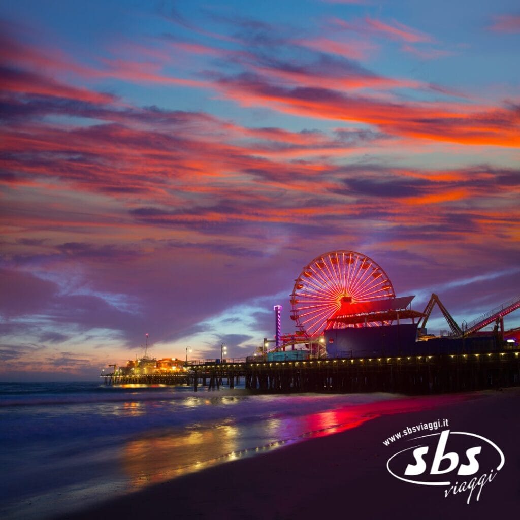 Una ruota panoramica su un molo di Los Angeles è illuminata al tramonto, con nuvole rosa e arancioni vivaci nel cielo e l'oceano in primo piano. Il logo di SBS Viaggi, ispirato a West 1924, appare nell'angolo.