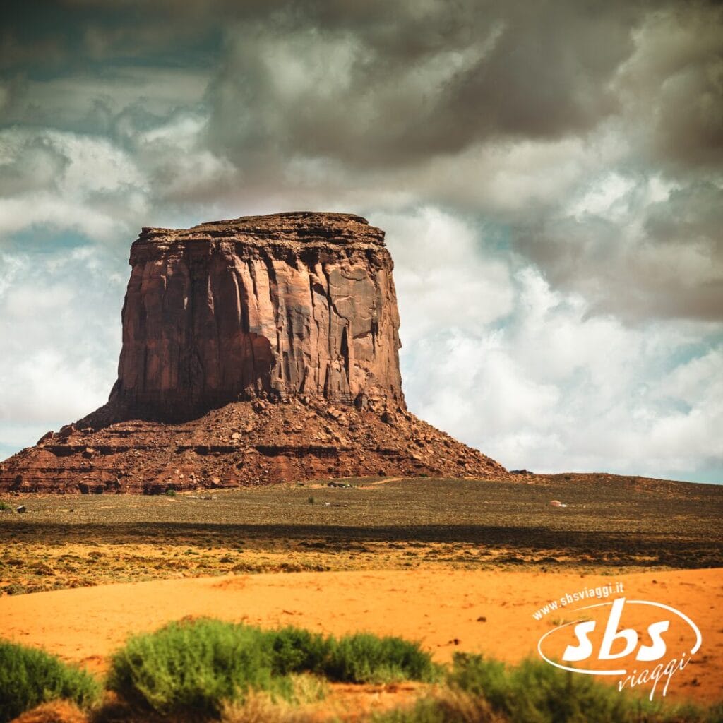 Un'ampia collina di arenaria si erge da un paesaggio desertico pianeggiante sotto un cielo nuvoloso; i verdi arbusti in primo piano evocano un'aura Essential del 1924. Il logo