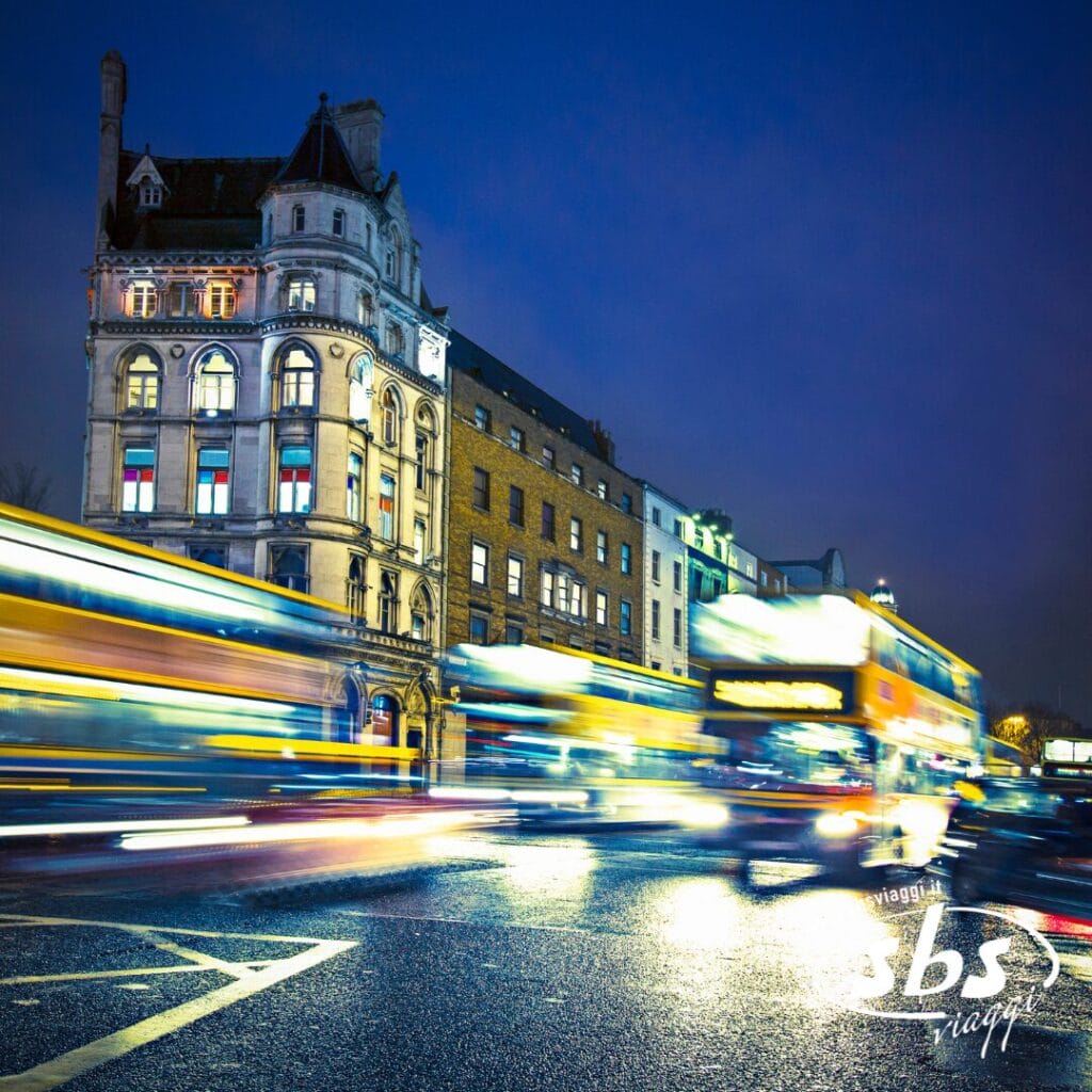 Una strada cittadina di notte con scie luminose prodotte da autobus e auto in movimento, edifici storici sullo sfondo e un accogliente bar dove la gente del posto sorseggia i classici drink irlandesi.
