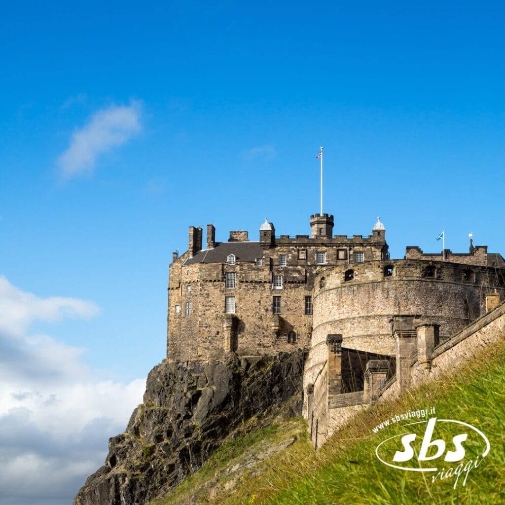 Un castello di pietra si erge sulla cima di una collina rocciosa sotto un cielo azzurro e limpido sull'isola di Scozia, con una bandiera che sventola dalla sua torre e un pendio erboso in primo piano, incarnando il fascino senza tempo dei castelli scozzesi.
