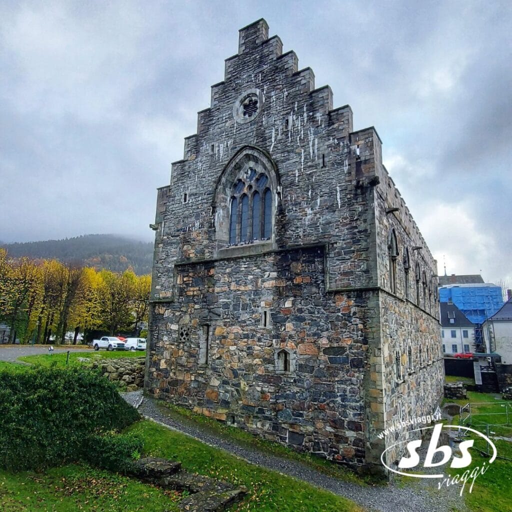Un edificio medievale in pietra con finestre ad arco e un timpano a gradini sorge su un'area erbosa in Norvegia, la Terra dei Fiordi, circondato da alberi e altri edifici sotto un cielo nuvoloso.