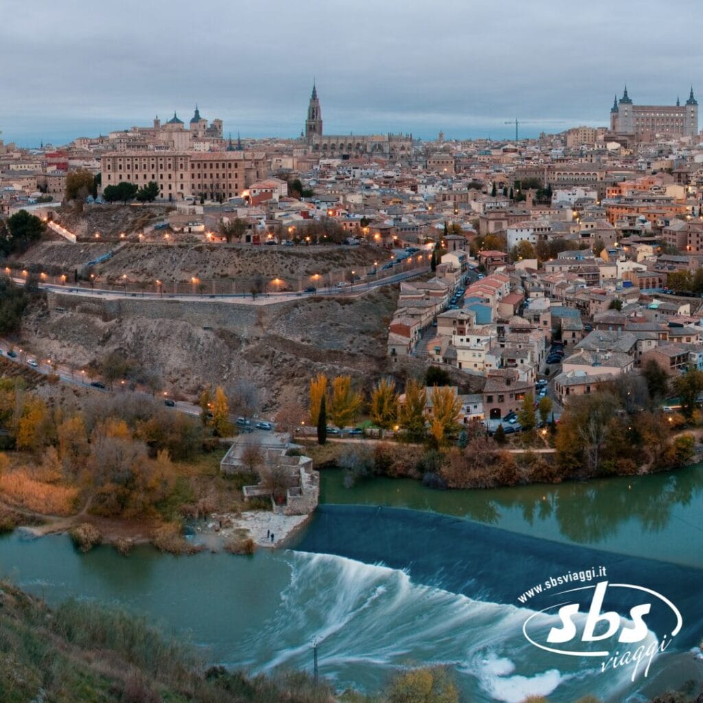 Vista aerea di una città storica che riflette il fascino della Spagna Medievale, con un fiume che scorre in primo piano. Lo skyline, ideale per un tour avvincente, presenta antichi edifici e un'imponente cattedrale sotto un cielo coperto.