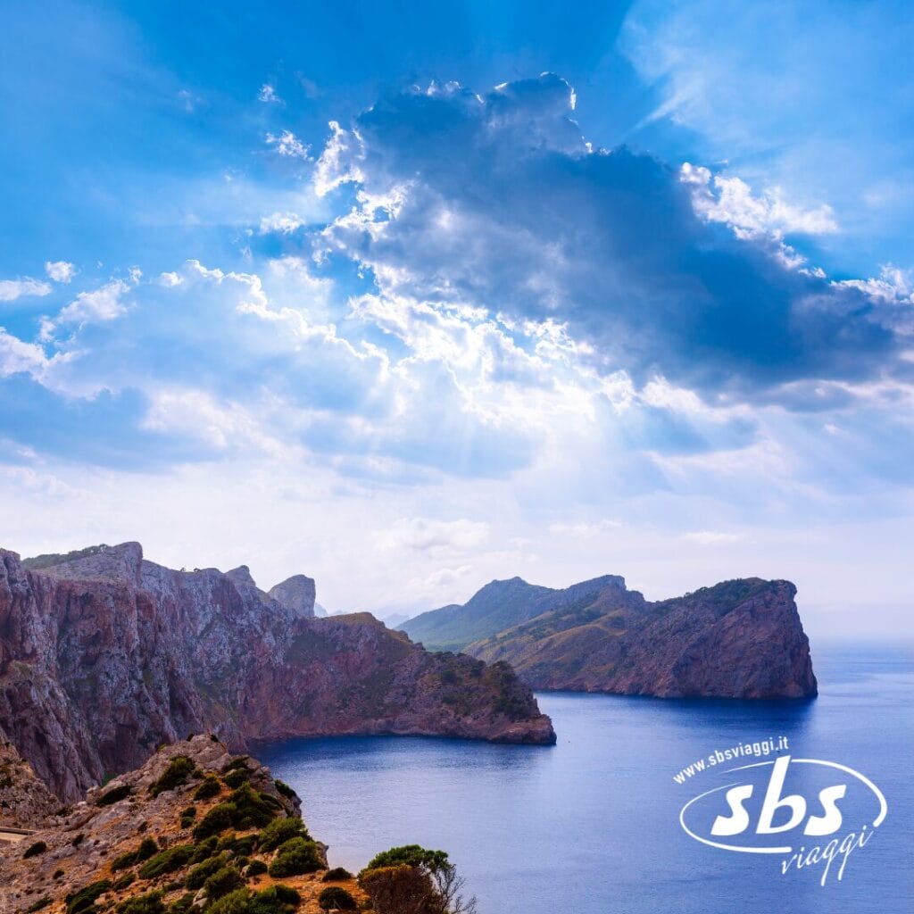 Costa rocciosa con ripide scogliere e mare azzurro sotto un cielo parzialmente nuvoloso, con raggi di sole che filtrano: uno scenario ideale per un tour a Capo Nord. Il logo