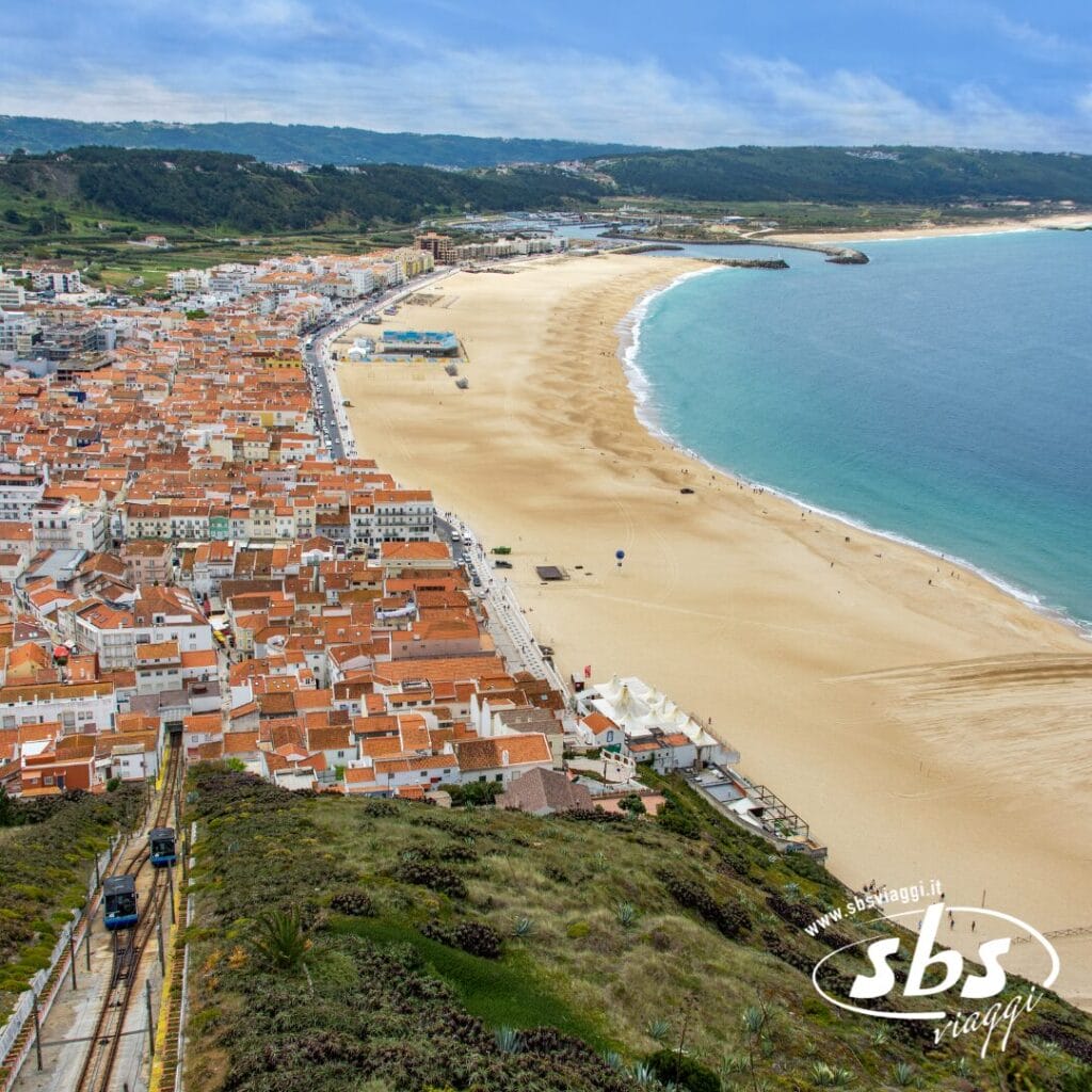 Vista aerea di una città costiera del Portogallo, con una lunga spiaggia sabbiosa che si estende lungo l'oceano. Edifici dai tetti rossi costeggiano la riva e verdi colline sono visibili sullo sfondo, rendendola una delle meraviglie che vale la pena esplorare durante il vostro tour.
