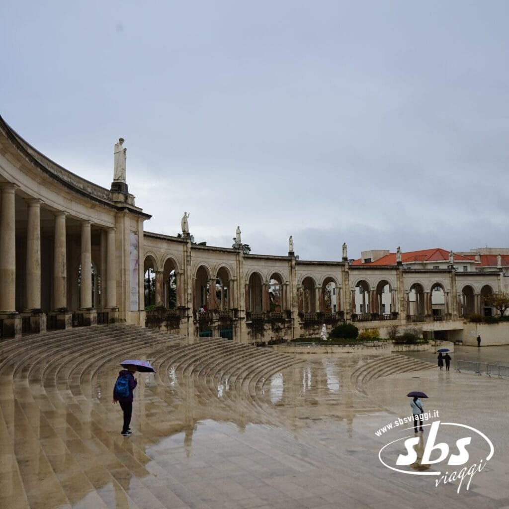Persone con ombrelli camminano in una piazza vuota e umida, con colonnati ad arco e statue, sotto un cielo nuvoloso, come se facessero parte di un tour improvvisato attraverso le pittoresche strade del Portogallo.