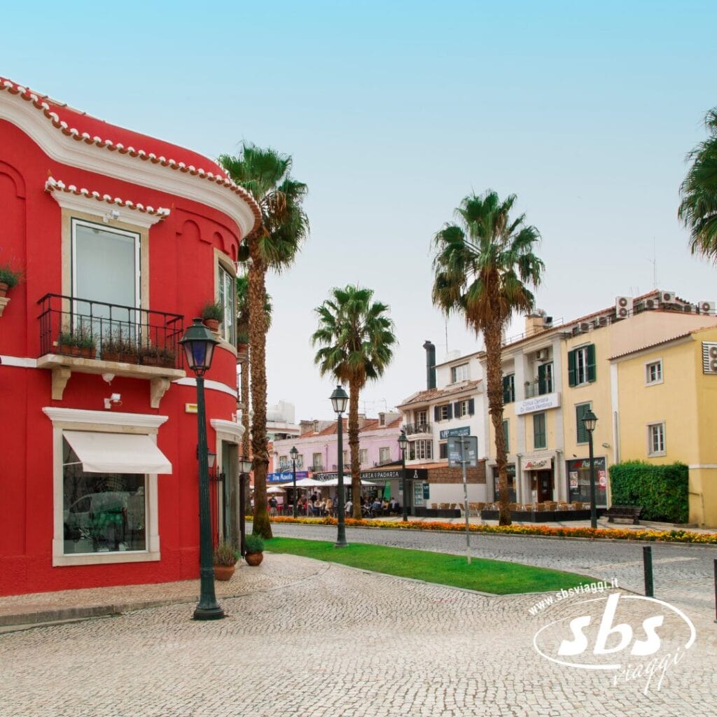 Una strada acciottolata con un sorprendente edificio rosso sulla sinistra, fiancheggiato da alte palme. Edifici colorati delineano lo sfondo, ricordando un paesaggio pittoresco direttamente dal Portogallo, il tutto sotto un cielo limpido che invita all'esplorazione.