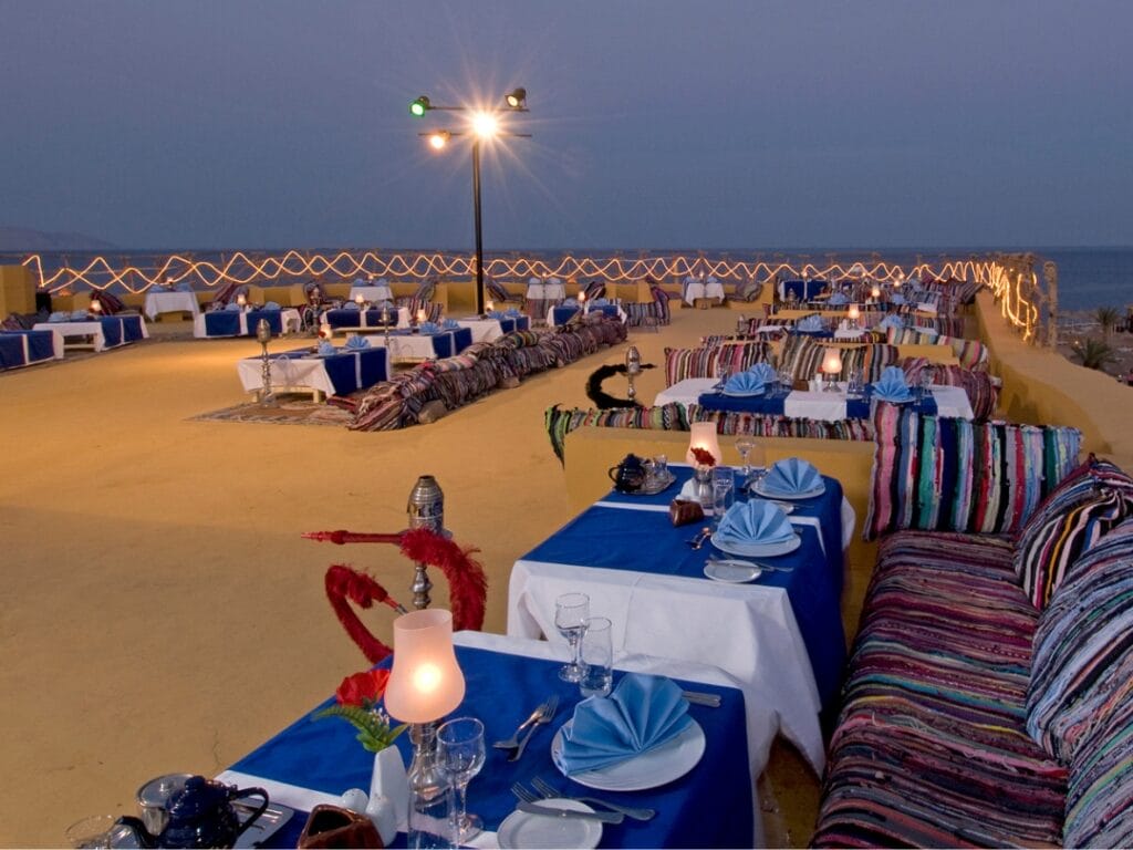 Al tramonto, questo ristorante all'aperto presenta tavoli con tovaglie blu e tovaglioli piegati con cura. Colorate sedute imbottite e luci a stringa Bozza automatica esaltano la vista del mare sullo sfondo, creando un'esperienza culinaria pittoresca.