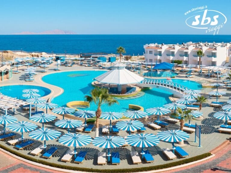 Il Dreams Beach Resort è un rifugio costiero a 4 stelle con una grande piscina, ombrelloni a strisce e lettini prendisole con vista sul mare. Edifici bianchi e una passerella si stagliano sullo sfondo sotto un cielo azzurro e limpido, offrendo la perfetta esperienza di resort sulla spiaggia.