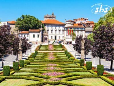 Giardino simmetrico con siepi curate, aiuole, statue e scalinate che conducono a edifici storici con tetti rossi in una piazza cittadina europea: un luogo ideale per un tour di Santiago attraverso l'affascinante Portogallo.