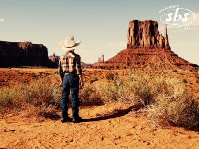 Un bambino con un cappello da cowboy è in piedi di fronte a una grande formazione rocciosa rossa in un paesaggio desertico, pronto a visitare gli Stati Uniti occidentali e ad ammirare gli straordinari parchi naturali degli Stati Uniti sotto un cielo limpido.