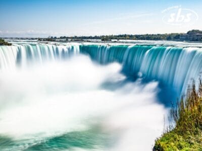 Un'ampia visuale delle Cascate del Niagara mostra l'acqua che precipita oltre il bordo, creando nebbia e correnti bianche, con vegetazione ed edifici di New York visibili sullo sfondo sotto un cielo limpido.