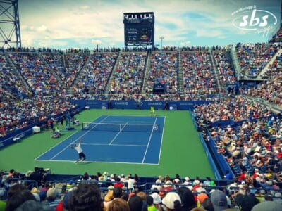 Una grande folla assiste a una partita di tennis professionistica su un campo in cemento blu e verde in uno stadio all'aperto, sotto un cielo parzialmente nuvoloso, durante il Tennis Grand Tour 2025.