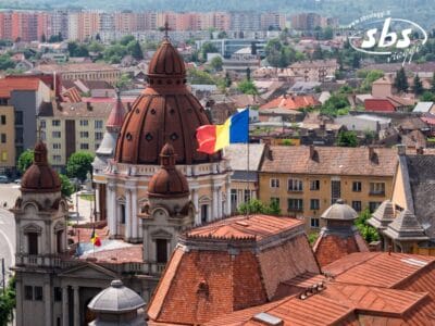 Un paesaggio urbano con un'imponente chiesa a cupola, la bandiera rumena che sventola e palazzi residenziali sullo sfondo sotto un cielo limpido invita a visitare il vivace cuore urbano della Romania.