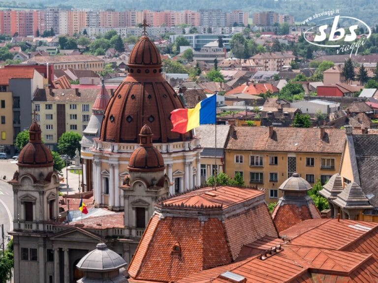 Un paesaggio urbano con un'imponente chiesa a cupola, la bandiera rumena che sventola e palazzi residenziali sullo sfondo sotto un cielo limpido invita a visitare il vivace cuore urbano della Romania.