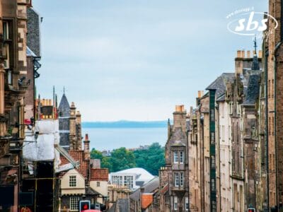 Vista lungo una stretta strada cittadina fiancheggiata da vecchi edifici in pietra in Scozia, che conduce a un lontano specchio d'acqua e vegetazione sotto un cielo nuvoloso: perfetto per chi cerca autentiche esperienze turistiche in Scozia.