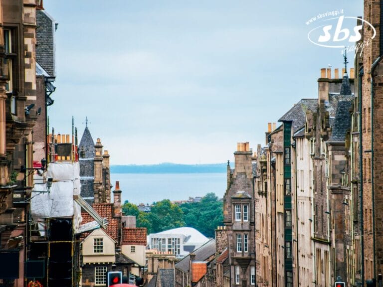 Vista lungo una stretta strada cittadina fiancheggiata da vecchi edifici in pietra in Scozia, che conduce a un lontano specchio d'acqua e vegetazione sotto un cielo nuvoloso: perfetto per chi cerca autentiche esperienze turistiche in Scozia.