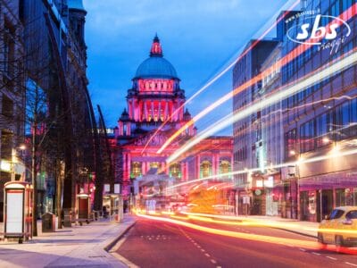 Strada cittadina al tramonto con le luci sfocate dei veicoli in transito; sullo sfondo lo storico edificio a cupola di Belfast in Irlanda illuminato da luci rosa e blu.