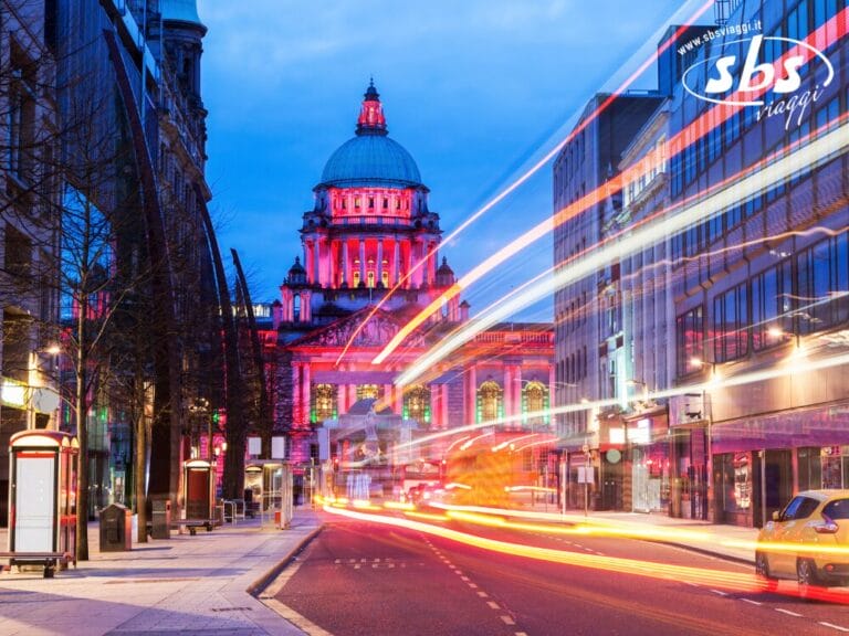 Strada cittadina al tramonto con le luci sfocate dei veicoli in transito; sullo sfondo lo storico edificio a cupola di Belfast in Irlanda illuminato da luci rosa e blu.