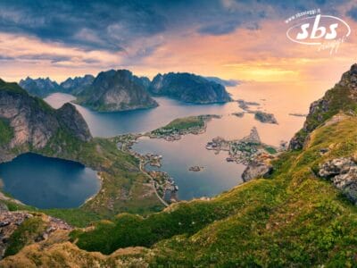 Una suggestiva vista aerea di un villaggio costiero incastonato tra montagne, laghi e mare al tramonto: un vero spettacolo dei Fiordi Norvegesi, con il logo di SBS Viaggi nell'angolo in alto a destra.