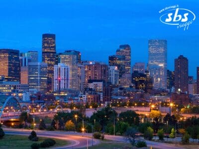Lo skyline della città di Denver al tramonto con edifici illuminati, un ponte in primo piano e il logo di SBS Viaggi nell'angolo in alto a destra: tutto ciò cattura lo spirito moderno di Colorado 1924.