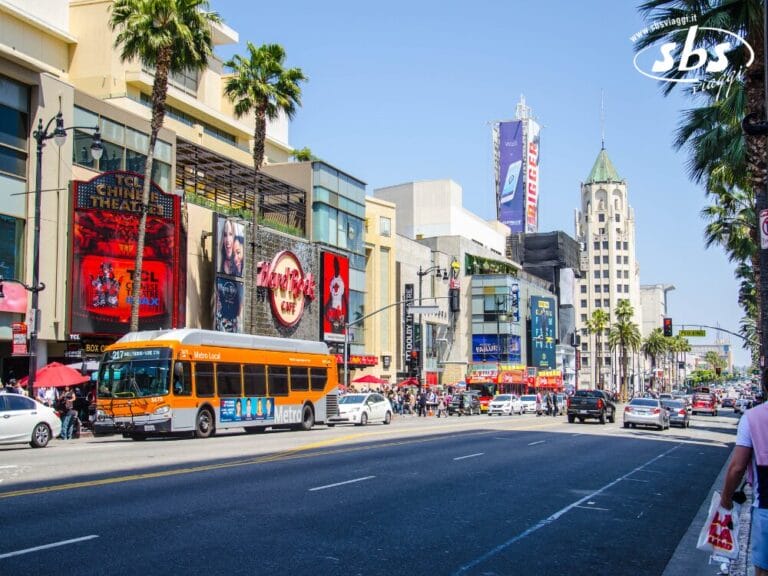 Una vista di una strada trafficata di Hollywood Boulevard a Los Angeles, con un autobus della metropolitana, pedoni, palme e tendoni dei teatri sotto un cielo limpido e luminoso: una scena che rispecchia davvero l'Essential 1924.