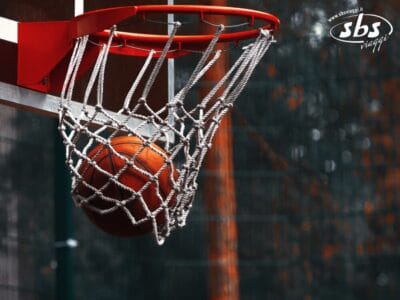 Un pallone da basket passa attraverso una rete bianca fissata a un canestro rosso in un campo all'aperto, evocando l'emozione di un'esperienza NBA lontano dal Madison Square Garden.