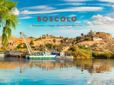 Una barca sul fiume a Il Cairo con edifici e palme su una collina sotto un cielo azzurro; la scritta "BOSCOLO" sopra, evoca la classica atmosfera della Crociera sul Nilo.