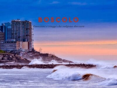 Le onde si infrangono su una costa rocciosa al tramonto, con alti edifici sullo sfondo; sopra il testo "BOSCOLO", che invita a visitare l'Argentina e a vivere momenti autentici.