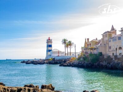 Vista panoramica sulla costa con un faro, palme ed edifici decorati in riva al mare sotto un cielo limpido, che ricorda una cartolina del Tour Portogallo.