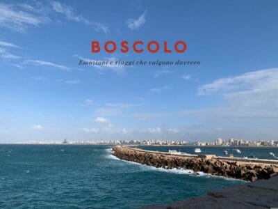 Un molo roccioso si estende nel mare azzurro sotto un cielo parzialmente nuvoloso, con in lontananza lo skyline di una città che ricorda il Cairo, lungo le rive del Nilo.