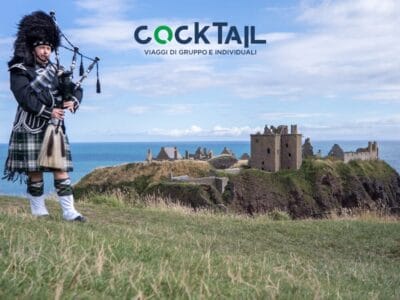 Un uomo in abiti tradizionali scozzesi suona la cornamusa vicino a una rovina di un castello costiero sotto un cielo azzurro, catturando lo spirito di un Tour Scozia attraverso Castelli e Isole.