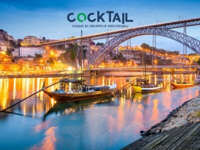 Barche tradizionali sul fiume Douro con le luci della città e un grande ponte ad arco a Porto, in Portogallo, al tramonto: lo scenario perfetto per un cocktail rilassante dopo il vostro Tour Portoghese.