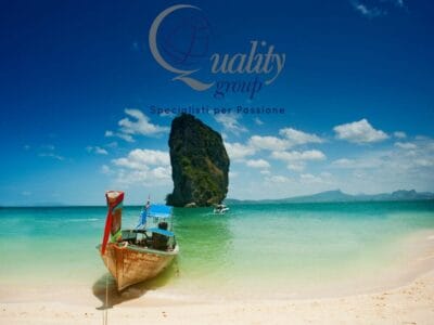 Una barca di legno galleggia vicino a una spiaggia sabbiosa con una grande isola rocciosa in acqua turchese sotto un cielo azzurro, catturando la serena bellezza di Koh Samui nella Thailandia del Nord.
