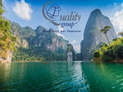 Lago panoramico con ripide scogliere verdeggianti; logo "Quality Group" e testo italiano sovrapposti al cielo, che vi invitano a scoprire Thailandia Experience 2025.
