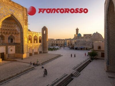 Piazza storica della città al tramonto, con persone che camminano e vanno in bicicletta lungo Via Della Seta e il logo "Francorosso" in rosso in alto.