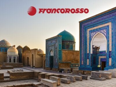 Antichi edifici con cupole turchesi al tramonto evocano la magia della Terra di Tamerlano, con il logo FRANCOROSSO nella parte superiore dell'immagine.