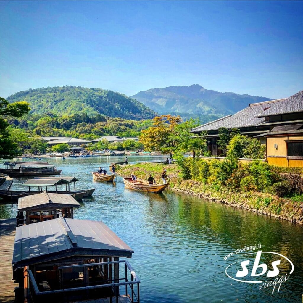 Barche di legno con passeggeri a bordo navigano lungo un fiume tranquillo in Giappone, passando accanto a edifici tradizionali e a lussureggianti colline verdi: un tour perfetto sotto un cielo azzurro e terso.