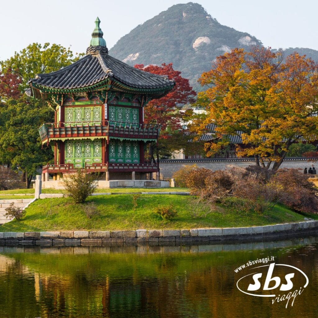 Un tradizionale padiglione coreano sorge su una piccola isola in uno stagno, circondato da alberi autunnali e da uno sfondo di montagne. L'immagine, perfetta per ispirarvi al Tour Corea del Sud, presenta il logo di sbsviaggi.