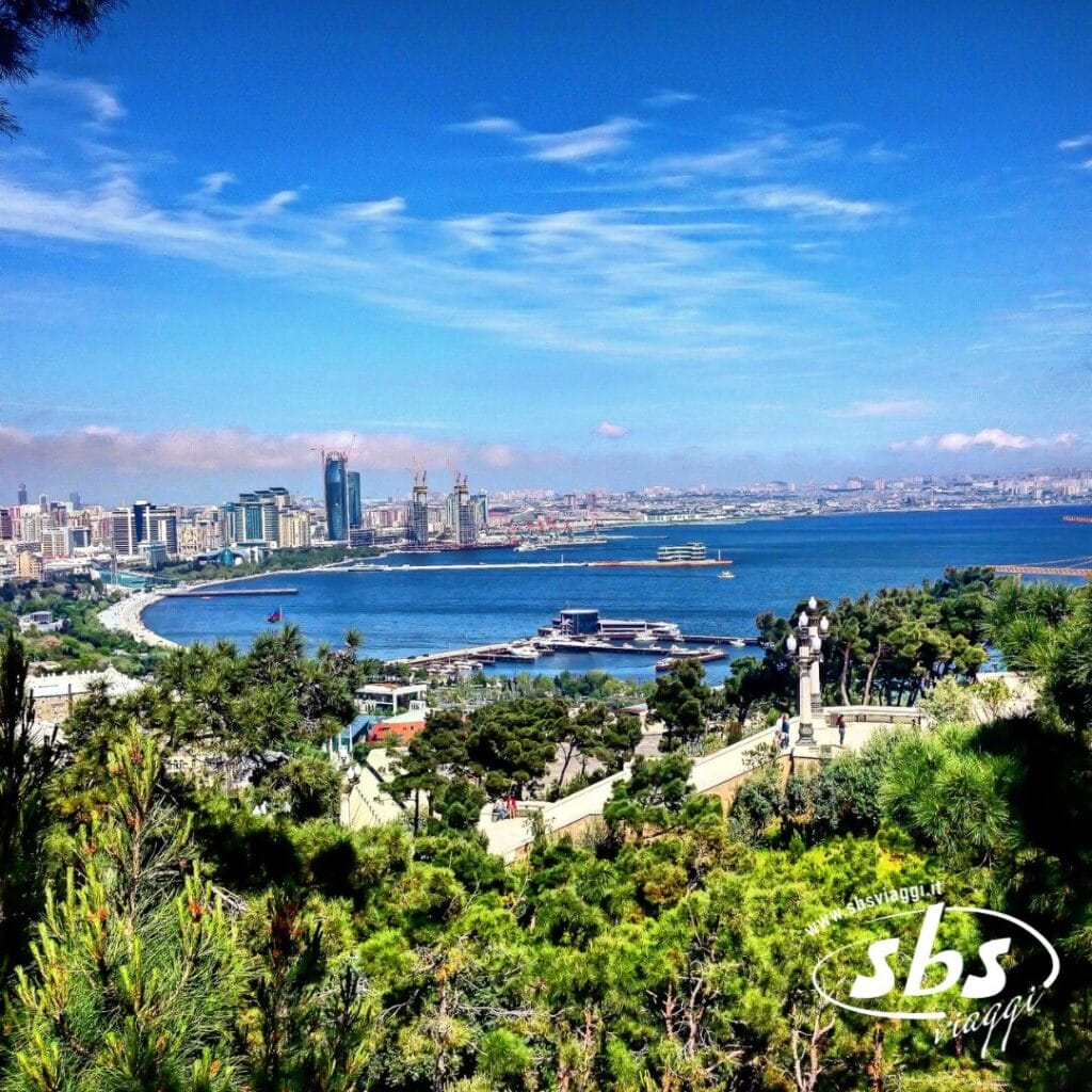 Una veduta urbana di Baku, in Azerbaijan, mette in risalto i moderni edifici lungo la costa del Mar Caspio, con vegetazione e alberi in primo piano sotto un cielo azzurro, a sottolineare la vibrante bellezza dell'Azerbaijan, vicino alla vicina Georgia.