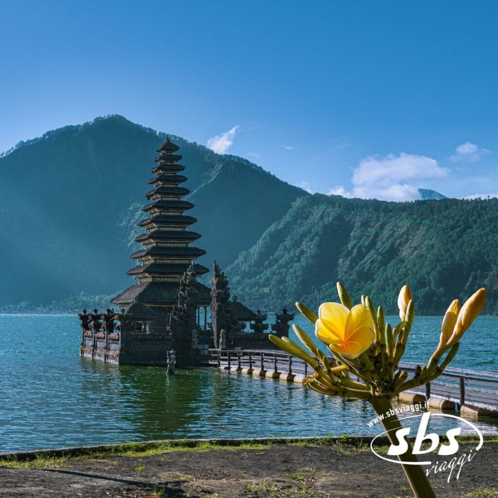Un tempio sul lago con tetti a più livelli si erge davanti alle verdi montagne di Bali, con un fiore giallo in primo piano e il cielo azzurro sopra: perfetto per il tuo prossimo post su Instagram. L'immagine presenta una filigrana