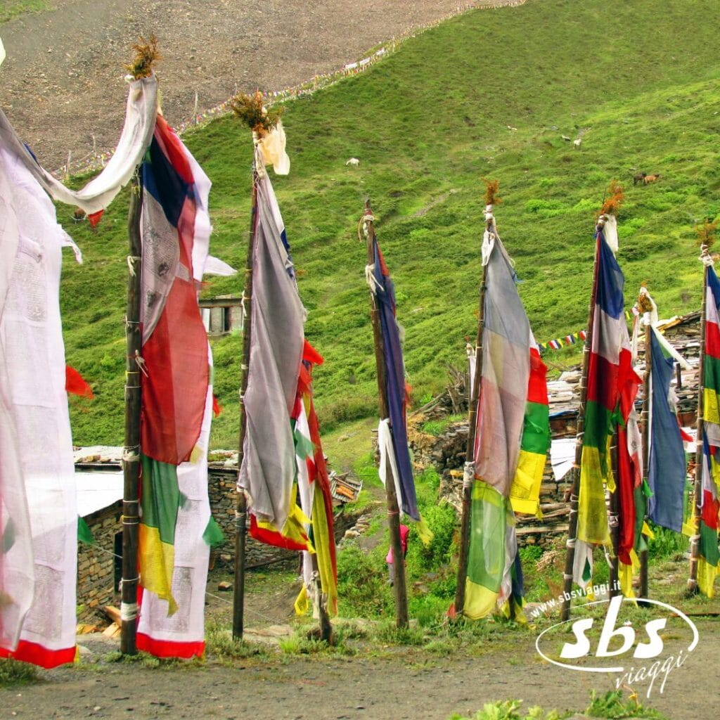 Bandiere di preghiera colorate su pali alti sono disposte in fila su un pendio erboso vicino alle strutture, con la Valle degli Dèi e le maestose montagne sullo sfondo: uno scenario perfetto per chi desidera visitare il Nepal.