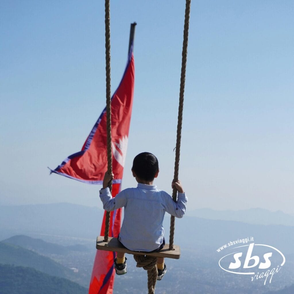 Un bambino con una camicia bianca è seduto su un'altalena di legno sospesa sopra le montagne del Nepal, tenendo in mano una grande bandiera rossa; nell'angolo appare il logo